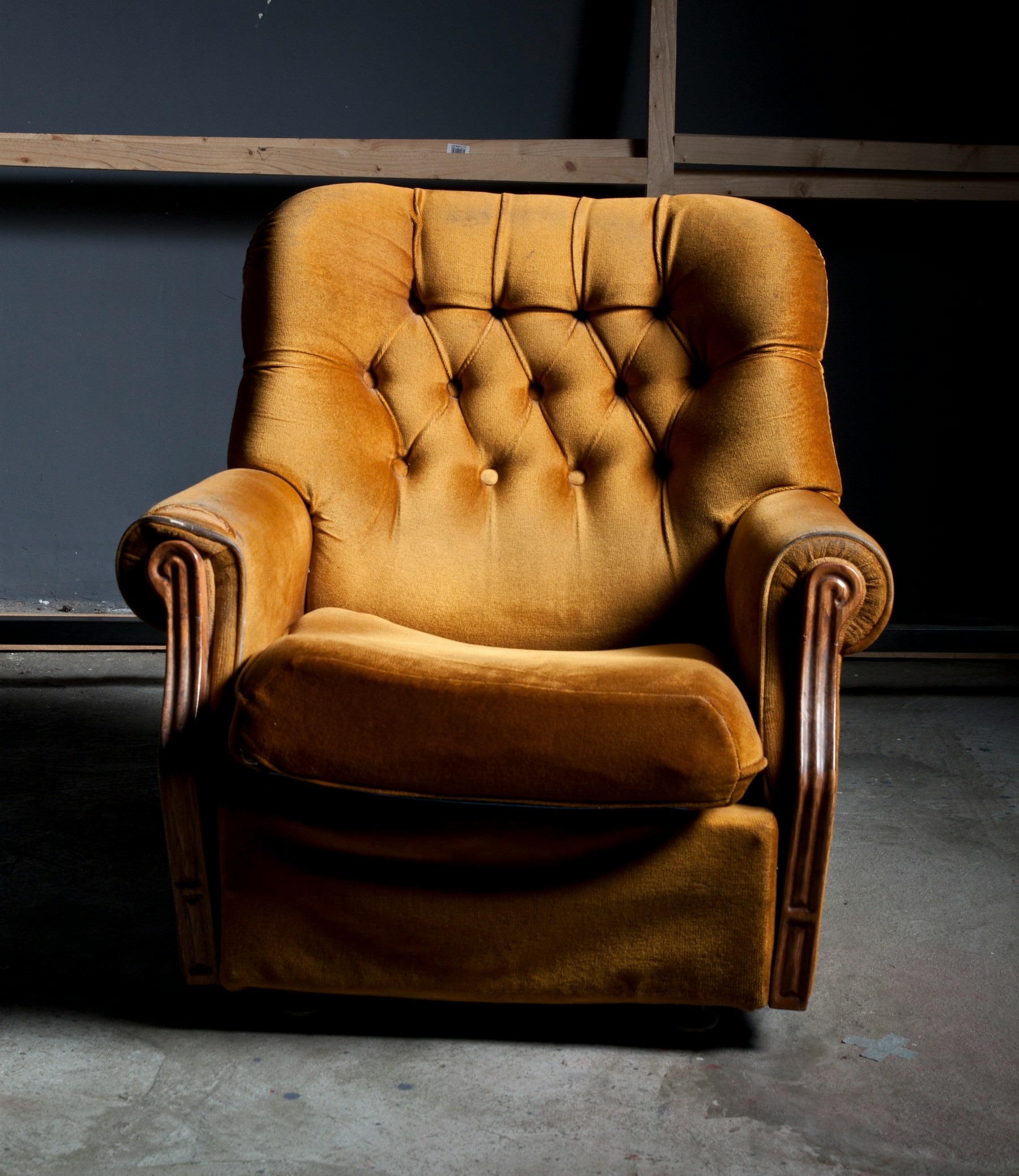 A button-tufted gold velvet armchair with scrolled wooden arms, standing alone against a bare workshop wall on a concrete floor.